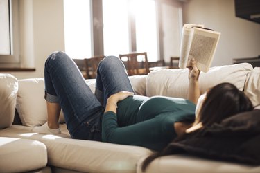 Pregnant person laying on couch reading a book, touching their stomach for fetal movement