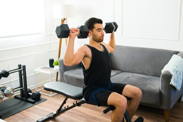 Man wearing black tank top and shorts doing a shoulder press on workout bench at home.