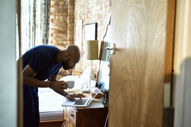 Man with coffee checking emails on laptop in bedroom