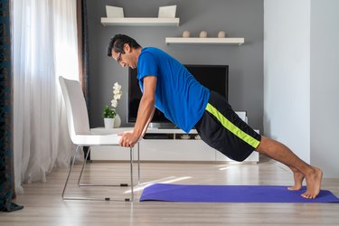an athlete wearing athletic shorts and a blue t-shirt does an incline push-up on a white chair in a modern living room on a purple yoga mat