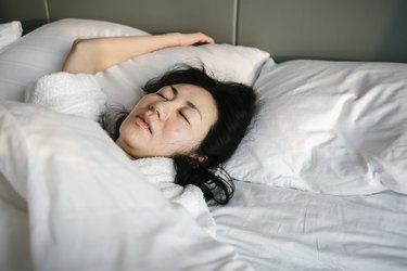 Person with long black hair sleeping under white bed sheets with their mouth slightly open, needing a nasal strip to breathe.