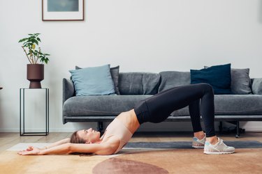 Person in beige sports bra and black leggings doing a Pilates shoulder bridge in living room to improve mobility.