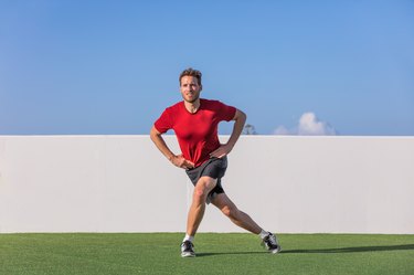 caucasian man in a red shirt doing a curtsy lunge on the grass in front of a blue sky