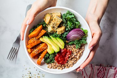 an overhead photo of hands holding a white bowl with quinoa, avocado, red onion, sweet potato and leafy greens on a marble table next to a fork