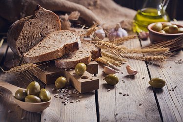 Fresh multigrain crusty bread, green olives and wheat ears on a rustic wooden table.