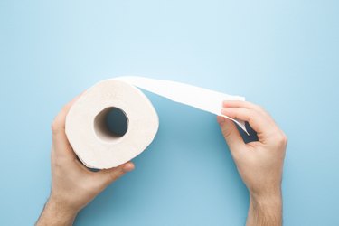Man's hands holding soft, white toilet paper roll on light pastel blue background, to represent feeling cold after pooping