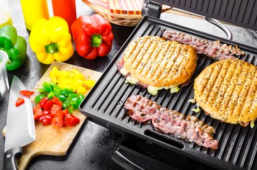 Panini maker with sandwiches and bacon, bell peppers on side on cutting board