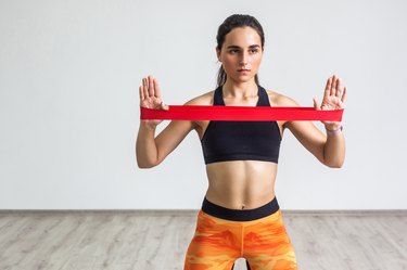 Portrait of young sporty woman wearing black top and orange leggings performs exercises for the muscles of the hands, workout with resistance band on white background.