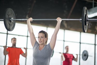 Person in gray T-shirt performing a military press with a barbell at the gym
