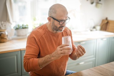Older person assigned male at birth in kitchen with glass of water and DHEA pill wondering about DHEA supplement mass review