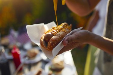 hand squeezing mustard over hotdog, one of the worst foods for longevity, outdoors