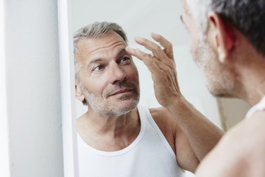 Person in white tank top looking in the mirror with a finger on their eyebrow, wondering how gray hair affects health