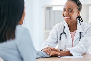 a smiling doctor wearing a white lab coat and stethoscope holds a patient's hand across a table