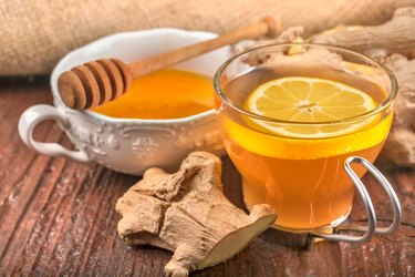 a glass mug of herbal ginger tea with a slice of lemon next to raw ginger and a honey bowl