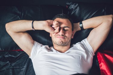 A man waking up in bed and rubbing his eyes because of eye discharge