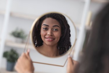 a woman smiling into a hand mirror, practicing body positivity