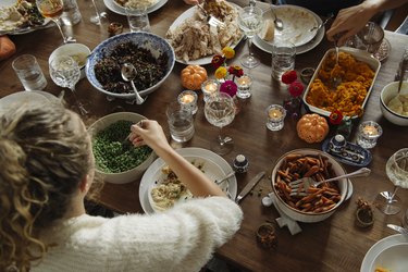 A overhead view of a person serving themselves a spoonful of peas at the Thanksgiving dinner table, surrounded by other side dishes.