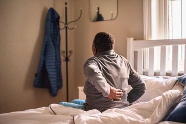 Person sitting up on the side of the bed with one hand on their back, suggesting pain after sleeping