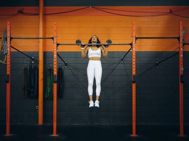 Person wearing a white sports bra and leggings doing a pull-up in a gym.