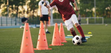 School boy running fast and kicking soccer ball on training slalom drill. Boy improving football skills on training. Coach observing pupil on training unit