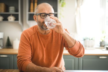 Man with glasses and orange sweater drinking glass of water in kitchen and wondering, "Can I drink salt water?"