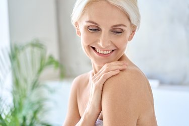 a smiling person with white hair wearing a bath towel and touching their bare shoulder