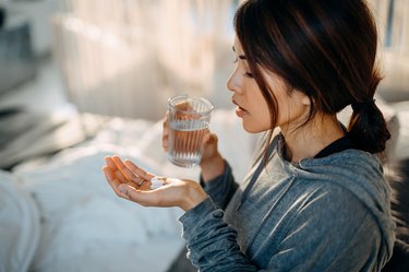 Young Asian woman sitting on bed and taking pain reliever pills, as a cold sore remedy