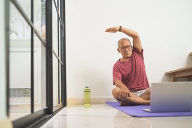 an older adult sits on a purple yoga mat in front of a laptop computer and next to a water bottle doing yoga for healthy aging