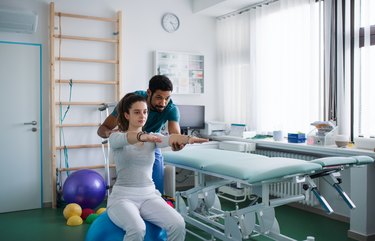 Physical therapist aiding patient sitting on blue yoga ball in room full of exercise equipment, as an example of pelvic floor physical therapy