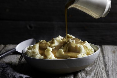 Jug of gravy being poured onto bowl of steaming mashed potatoes, studio shot