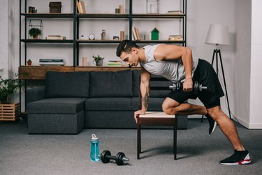 strong bi-racial man doing exercise with dumbbell on chair in living room