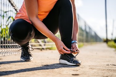 person tying shoelaces in a squat position wearing black leggings and an orange tank top before walking to build muscle