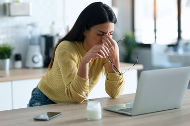 Stressed business woman working from home on laptop looking worried, tired and overwhelmed while sitting in the kitchen at home.