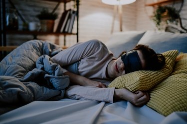 a person with brown hair in a bun sleeping in bed and wearing a black eye mask