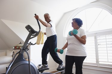 Smiling man using elliptical cardio machine at home with woman using hand weights