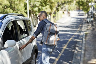 a person with a shaved head wearing jeans and a backpack opening the driver side door of a white car on the roadside
