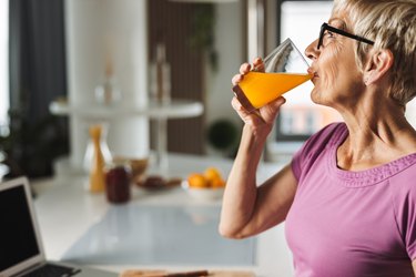 Older person assigned female at birth in purple shirt drinking fresh juice in the kitchen wondering why does orange juice make me vomit