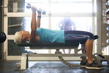Older woman lifting dumbbells while lying on her back on a bench in gym