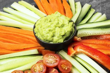 Guacamole in a bowl and various vegetables (carrots, tomatoes, cucumbers, celery) on a dark background