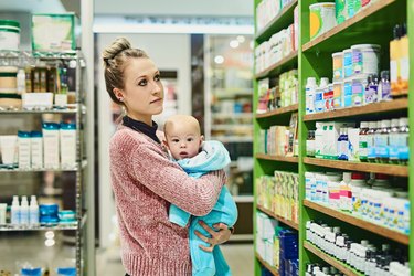 A person with blonde hair in a bun holding their baby in the drugstore and looking for a postnatal vitamin