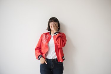 a person wearing a red jacket, white t shirt and black pants covers their face while laughing and blushing against a white wall