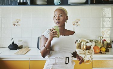 Closeup of person with short blonde hair in a white outfit sipping a green smoothie for constipation in their kitchen