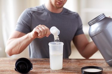 close up of a man with a grey shirt, pouring creatine powdered nutritional supplement into a clear protein shaker bottle, while holding the tub of powder