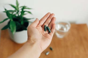 a close up of a hand holding ginkgo biloba dietary supplements in front of a small houseplant in a white pot and a glass of water