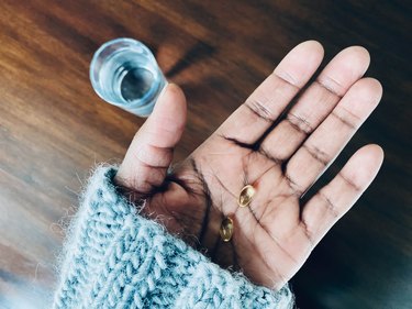 overhead shot of hand holding vitamin D capsules with glass of water on wooden table in the background