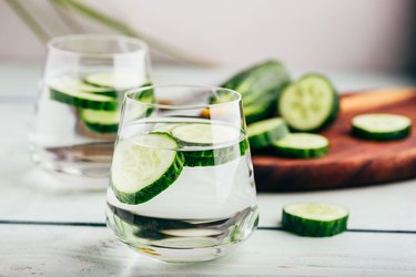 two glasses of cucumber water for health benefits like hydration with sliced cucumber on wooden cutting board