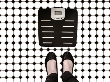 Body weight scales on tiled bathroom floor with woman's feet beside it like shes going to step on them - top view