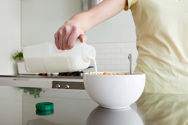 a close up of a person pouring a jug of milk into a large white bowl in a white kitchen