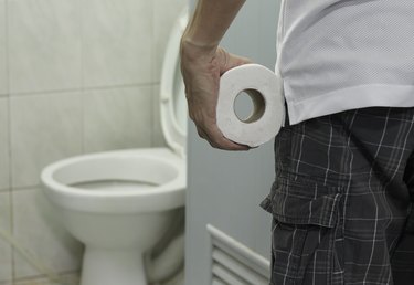 A close up of a person standing in the bathroom holding a roll of toilet paper in front of a toilet with the seat up