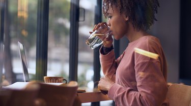 A woman sitting at a desk working on a laptop and drinking water for constipation relief